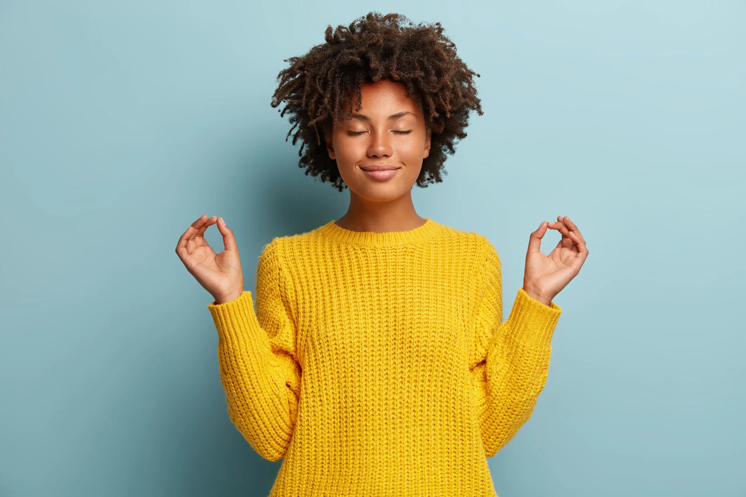 Satisfied dark skinned young woman makes okay gesture with both hands, keeps eyes closed, dressed in yellow clothes, practices yoga after work