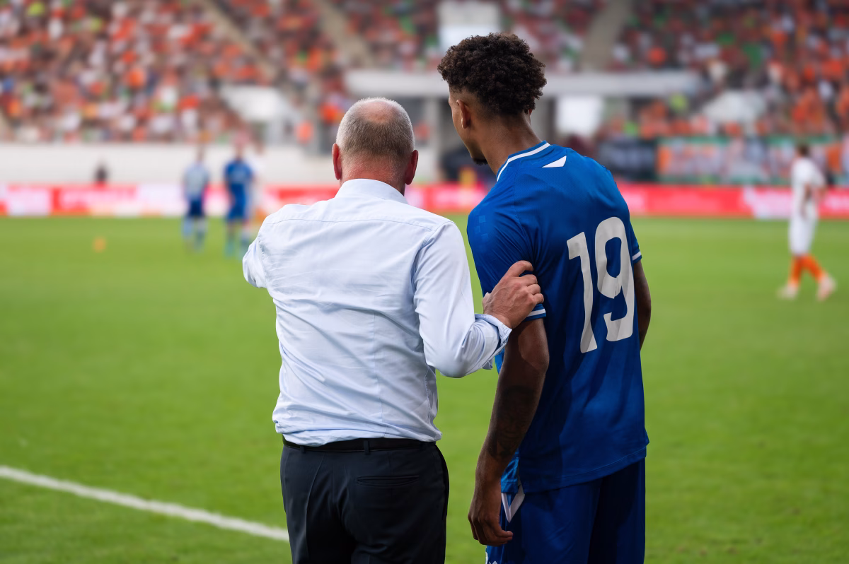 Coach discussing ongoing football/soccer game with an experienced player on the sideline in a stadium. 